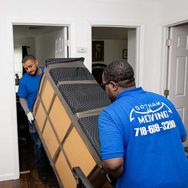 Two Gotham movers carrying a tall pad-wrapped armoire through an interior doorway