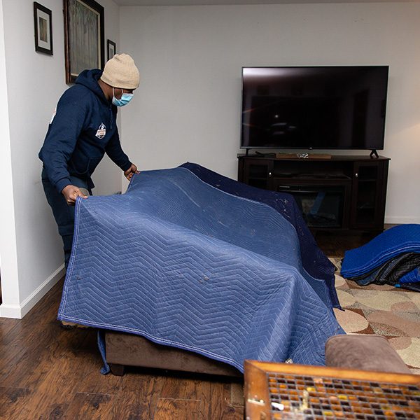 A mover laying a moving blanket over a sofa in a living room