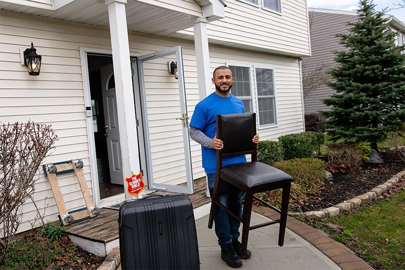 A Gotham mover in a blue shirt carrying a dining chair out of a house front door, a suitcase and dolly beside him