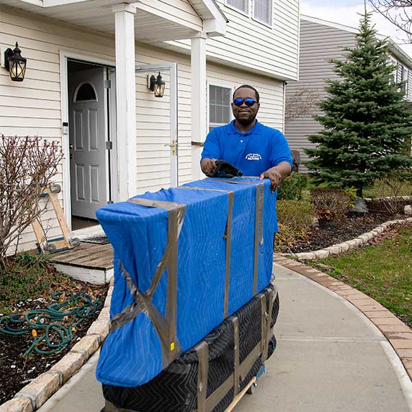 A Gotham mover wheeling pad-wrapped furniture on a dolly down a front walk