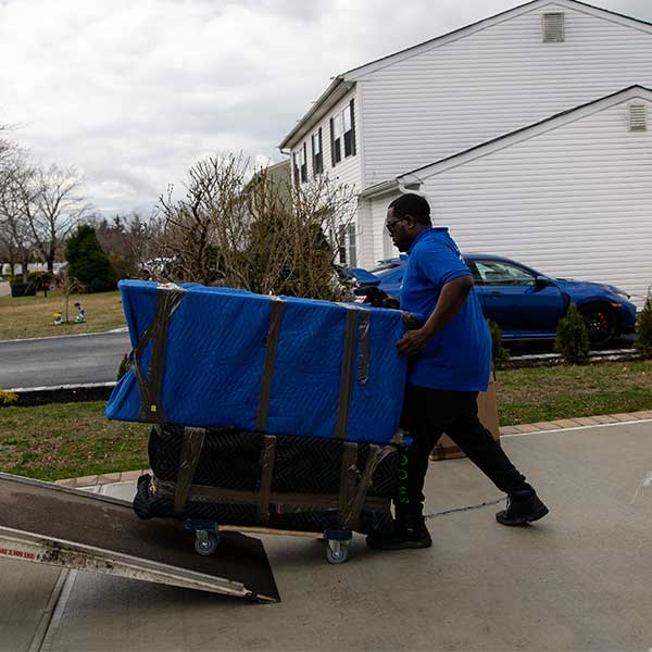 A Gotham mover pushing pad-wrapped furniture on a dolly off a truck ramp in a driveway