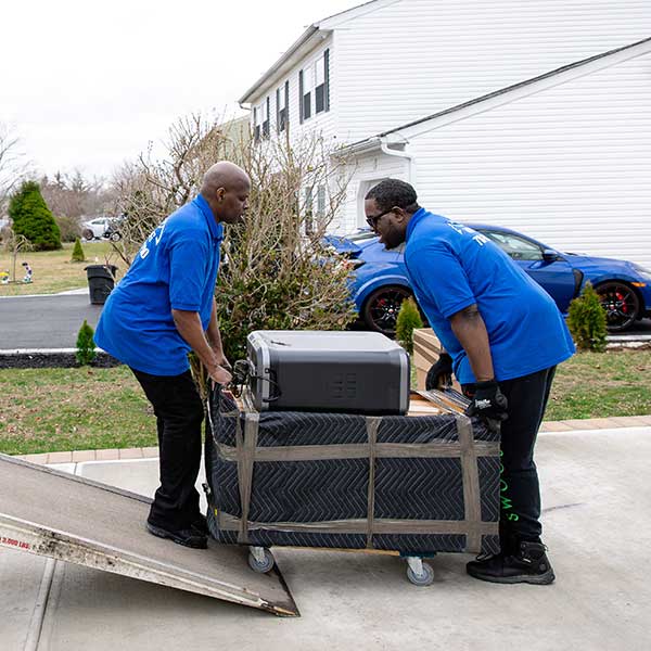Two Gotham movers rolling a pad-wrapped dresser on a dolly up a truck ramp in a driveway