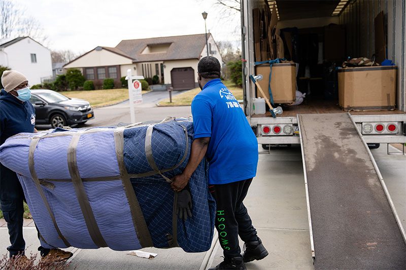 Two movers carrying a pad-wrapped piece toward a truck ramp, a house and driveway behind