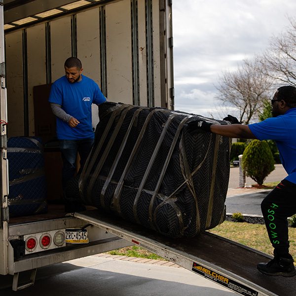 Two Gotham movers loading a pad-wrapped piece up the ramp into a box truck
