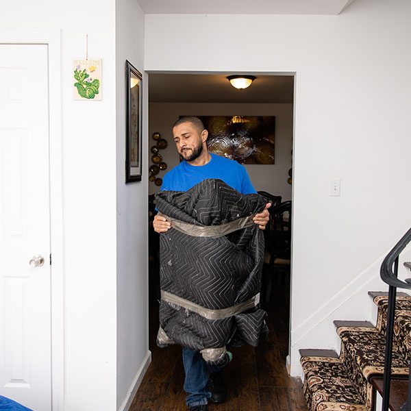 A Gotham mover carrying a pad-wrapped item down a hallway past a carpeted staircase