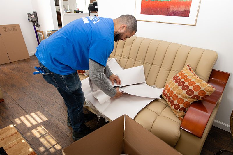 A Gotham mover wrapping a leather sofa in packing paper, a painting above