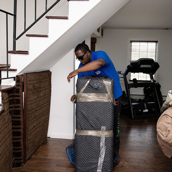A Gotham mover shrink wrapping a pad-wrapped item beside an indoor staircase