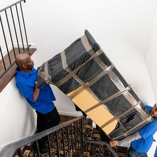 Two Gotham movers angling a pad-wrapped sofa up a narrow stairwell