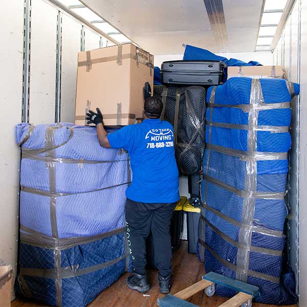 A Gotham mover lifting a carton onto a wall of pad-wrapped furniture inside a truck