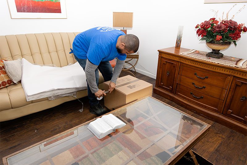 A Gotham mover taping a carton in a living room, sofa and cabinet protected in paper