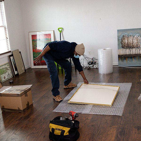 A mover setting a framed canvas face down on a sheet of bubble wrap, paintings leaning against the walls, a bubble wrap roll and tool bag nearby