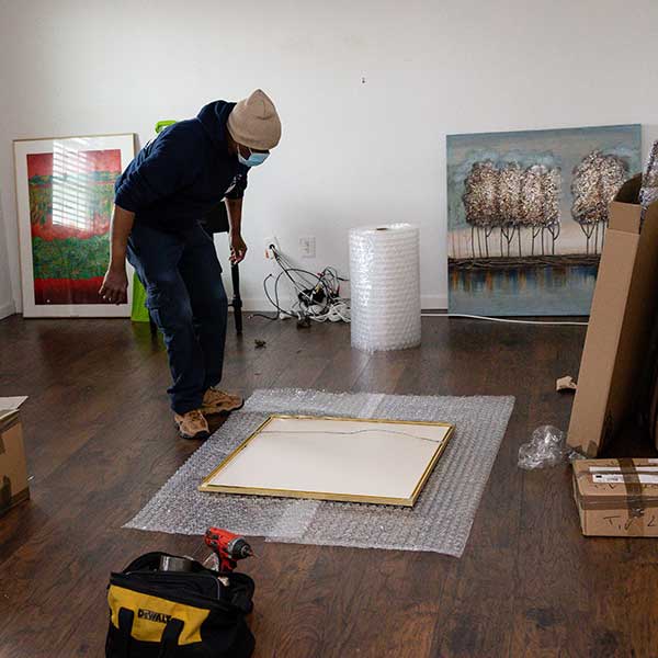 A mover bending over a framed piece laid on bubble wrap, large paintings leaning on the walls